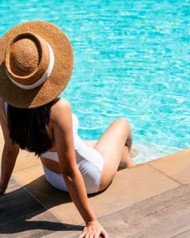 Woman in straw hat sitting on pool deck overlooking bright turquoise swimming pool