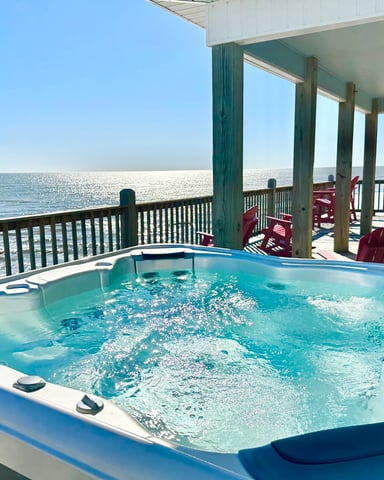 Hot tub on oceanfront deck with red Adirondack chairs and wooden railing overlooking calm blue water