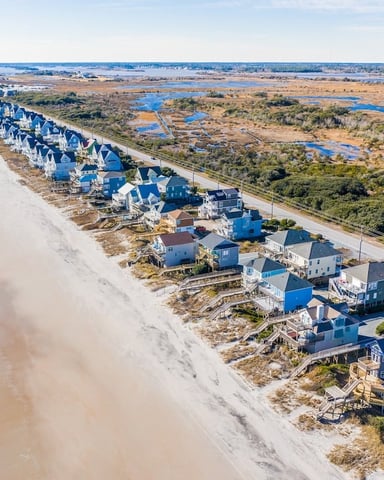 Aerial view of beachfront homes with blue roofs along sandy shore, ocean waves, and salt marshes inland
