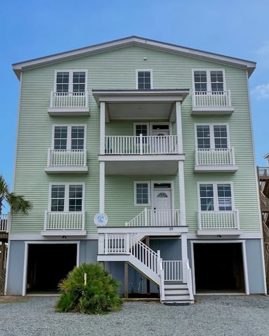 Three-story green beach house with white railings and multiple decks, flanked by coral and blue vacation homes.