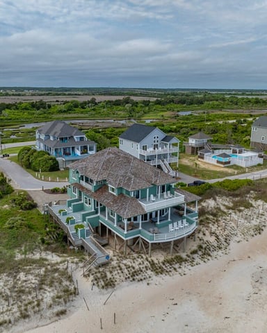 Aerial view of beachfront homes on stilts with dunes, marsh, and bay in background