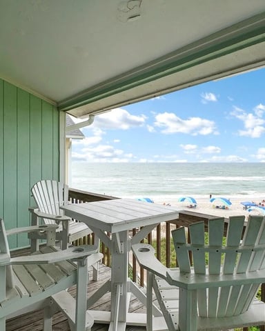 Covered beach house deck with white Adirondack chairs overlooking sandy beach and ocean with blue umbrellas