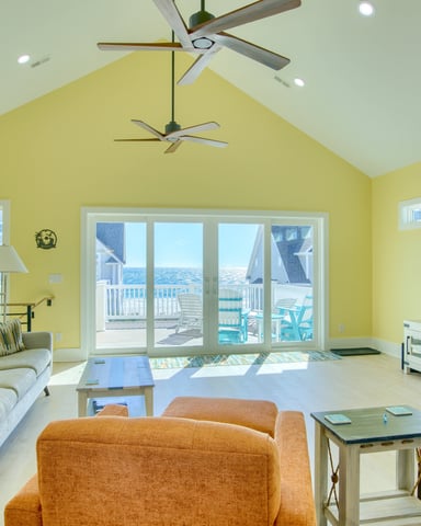 Bright yellow vaulted living room with ocean view, beige sofa, orange armchair, wall-mounted TV, and glass doors opening to deck