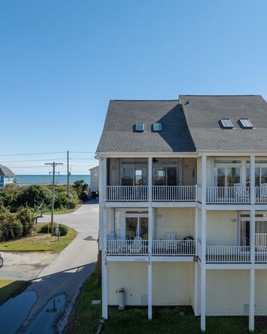 Beachfront house with gray roof, white railings, and multiple decks overlooking ocean, neighboring colorful beach homes visible