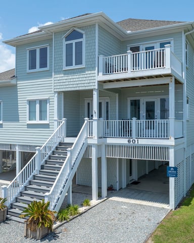 Light blue elevated beach house with white railings, multiple decks, and water view in background