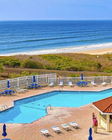 Oceanfront resort with swimming pools, white railings, blue lounge chairs, sandy beach, and dune vegetation beyond