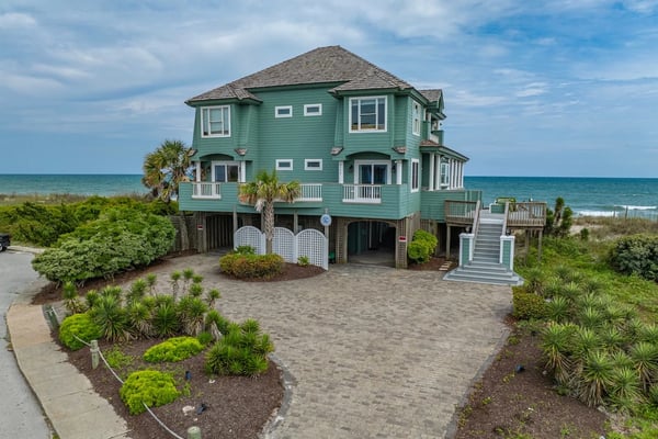 Turquoise beach house with elevated foundation, multiple decks, and ocean view behind palm trees and landscaping.