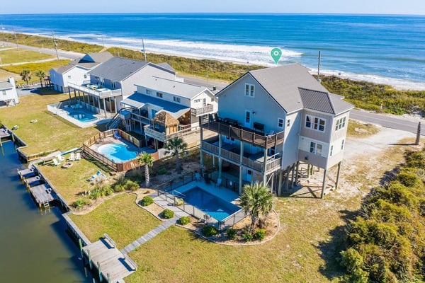 Aerial view of beachfront homes with pools, docks, and ocean beyond sandy dunes